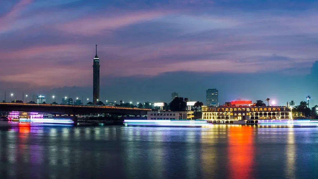 Cairo skyline with illuminated dinner cruise boat on the Nile River at dusk.