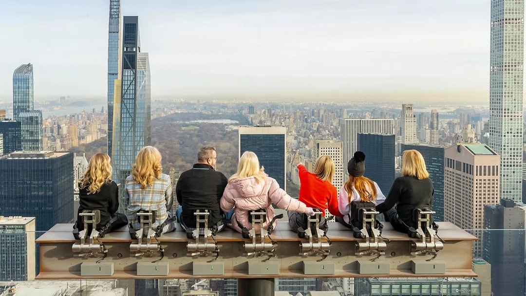 People sitting on The Beam at Top of the Rock, overlooking New York City skyline.