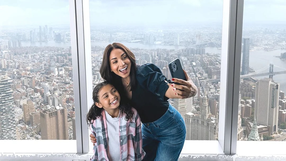 Visitors taking a selfie at One World Observatory with New York City skyline in the background.