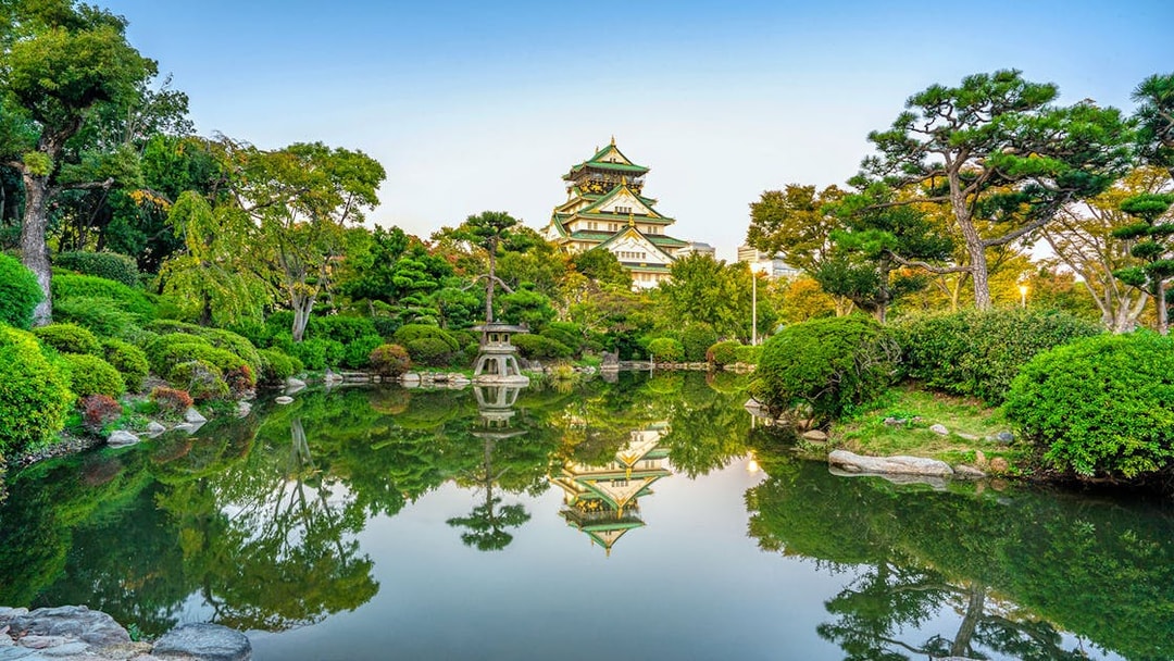 Osaka Castle reflected in a serene garden pond, surrounded by lush greenery.