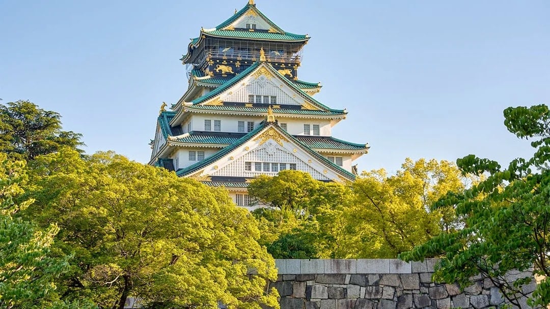 Osaka Castle exterior with lush green trees and blue sky.