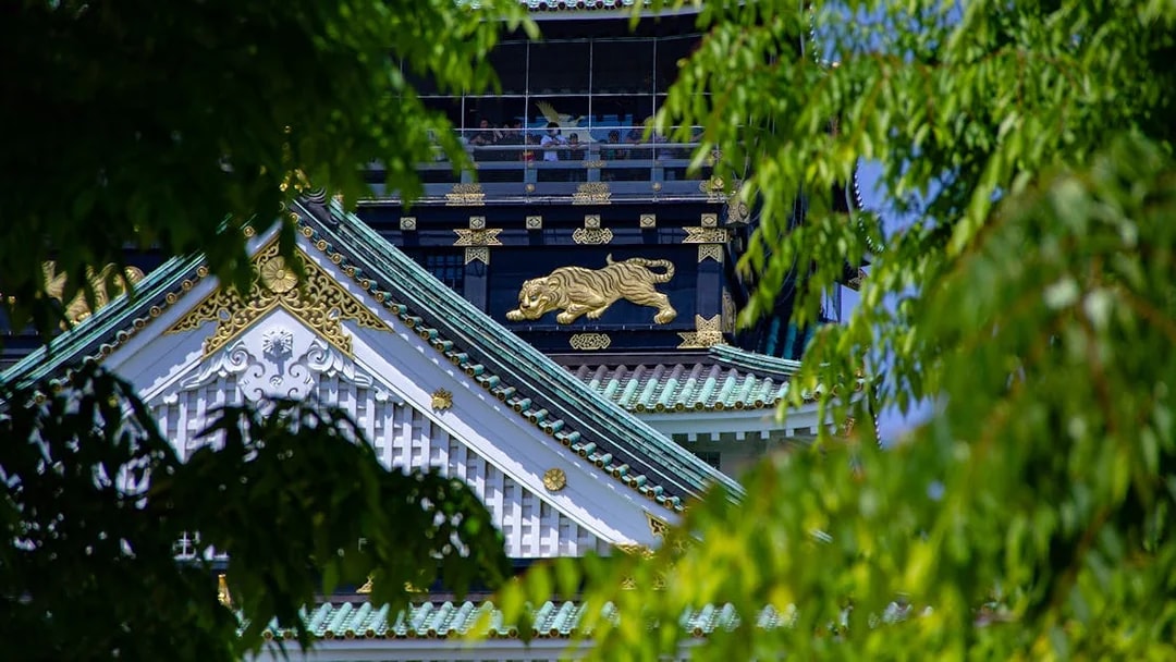 Osaka Castle rooftop with ornate gold tiger detail, partially framed by green leaves.