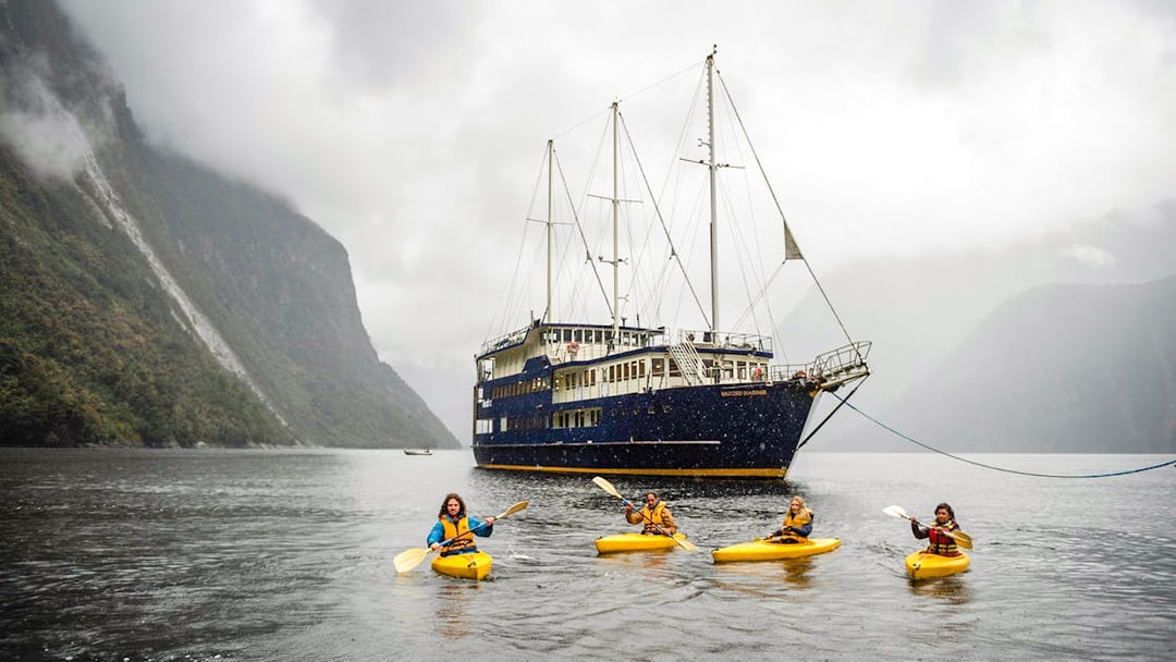 Kayakers paddle near a cruise ship in Milford Sound, New Zealand, surrounded by misty mountains.
