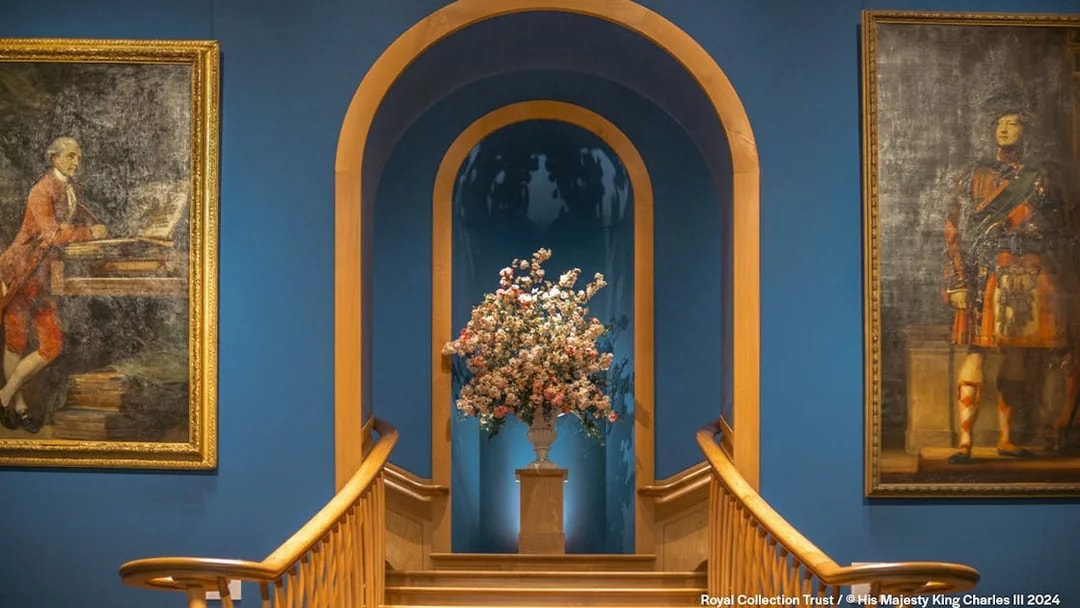 Floral arrangement on pedestal between two historical paintings in a blue-walled gallery.