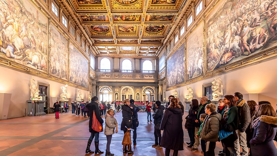 Visitors inside the ornate hall of Palazzo Vecchio, Florence, viewing large frescoes and sculptures.