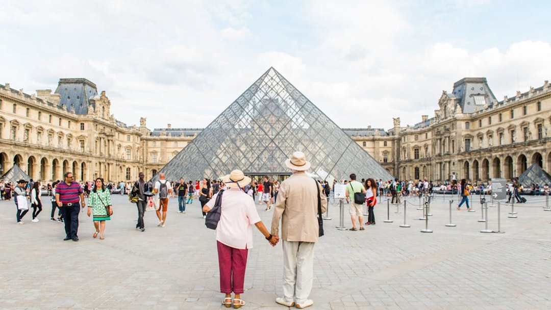 Old couple holding hands in front of the Louvre Pyramid, Paris.