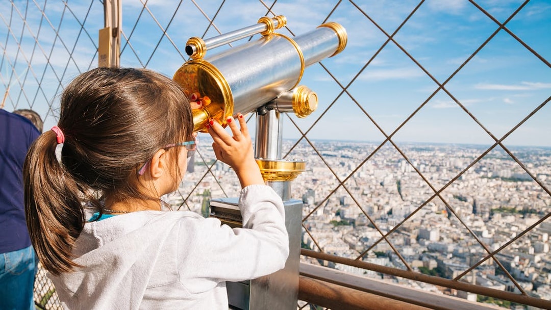 Child using telescope on Eiffel Tower viewing platform, Paris cityscape in background.