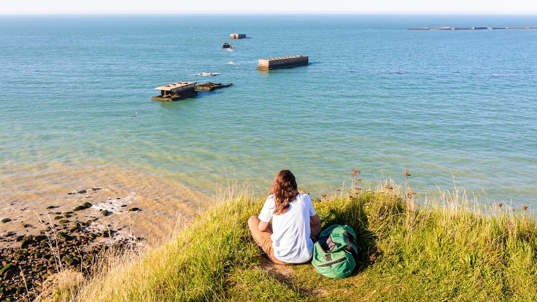 Person sitting on a grassy cliff overlooking the Normandy D-Day beaches with WWII remnants in the sea.