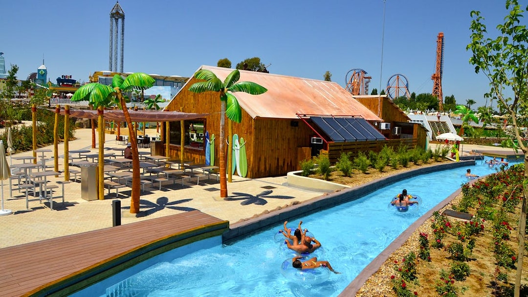 Lazy river with people on tubes at Parque Warner Beach, Madrid, with roller coasters in the background.