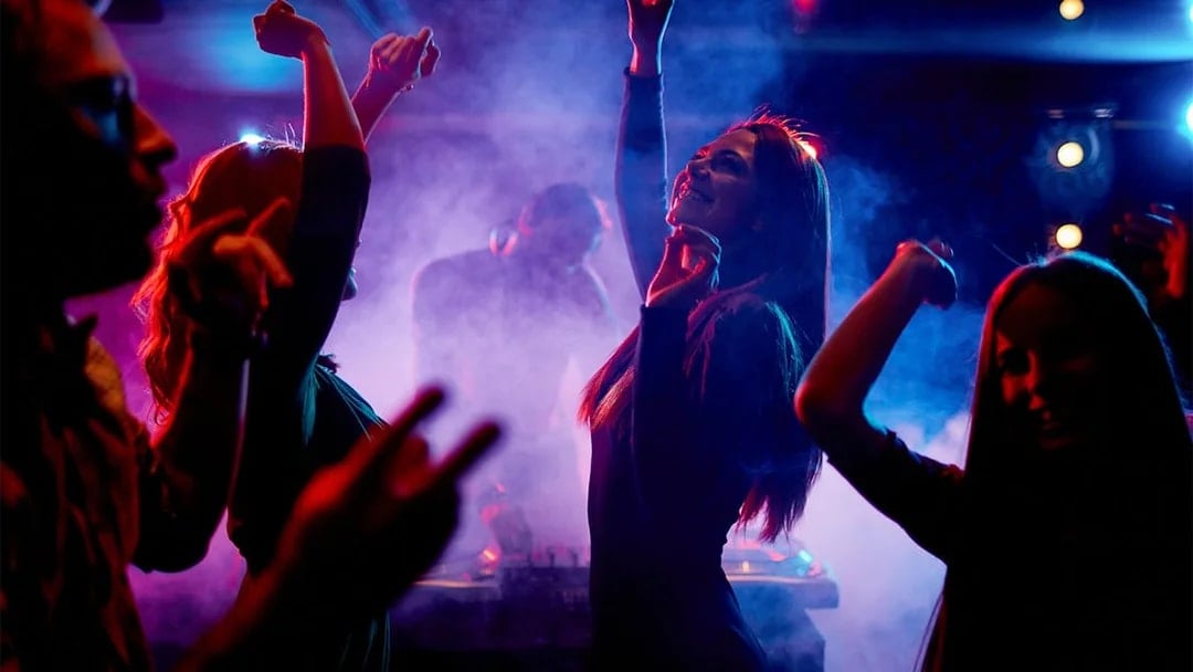 Women dancing energetically at a nightclub with a DJ in the background.