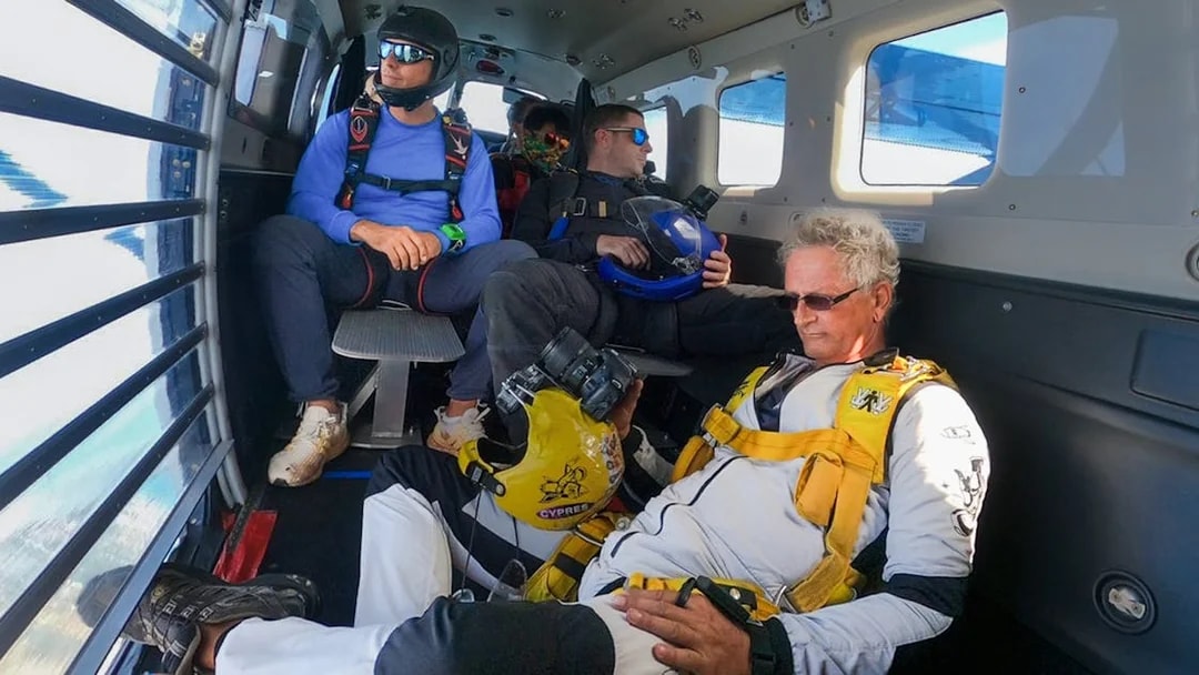 Tourists seated inside a skydiving plane wearing safety gear.