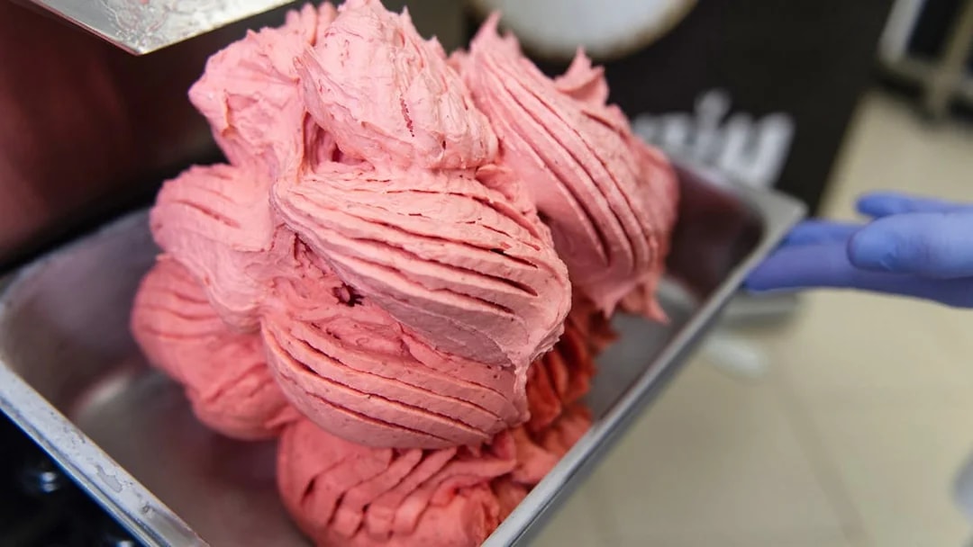 Freshly made pink gelato being scooped into a metal container.