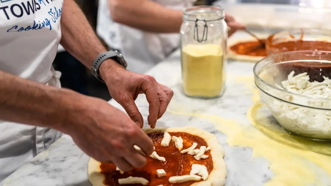 Hands preparing pizza dough with tomato sauce and cheese during a cooking class.