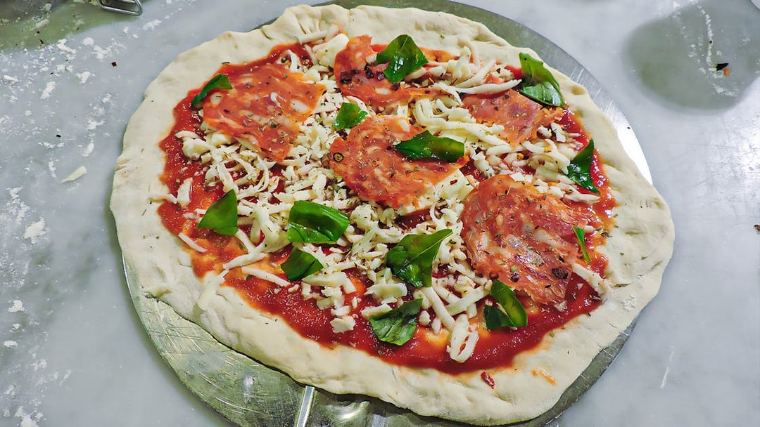 Pizza dough topped with tomato sauce, cheese, pepperoni, and basil during a cooking class.