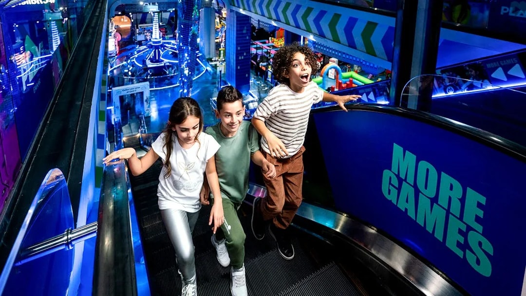 Children on escalator at Play DXB, Dubai, with arcade games in background.