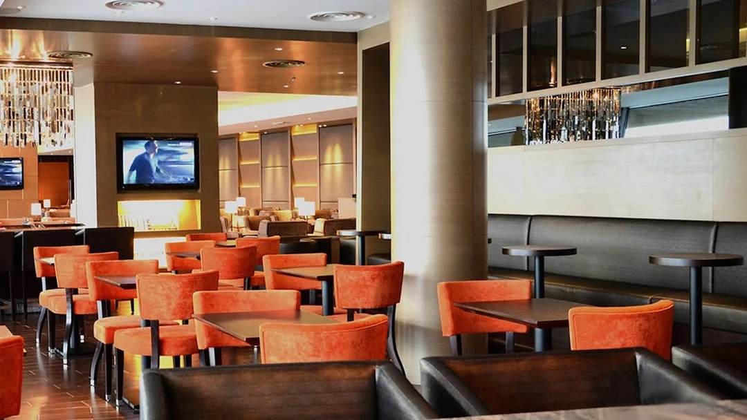 Plaza Premium First lounge seating area with orange chairs and a TV, Satellite Terminal.