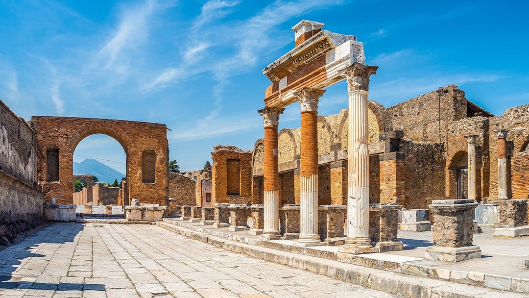 Ancient ruins of Pompeii with columns and archway, Mount Vesuvius in background.