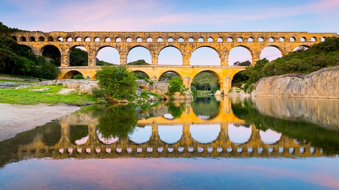 Pont du Gard aqueduct reflecting in the Gardon River, France, with surrounding greenery.