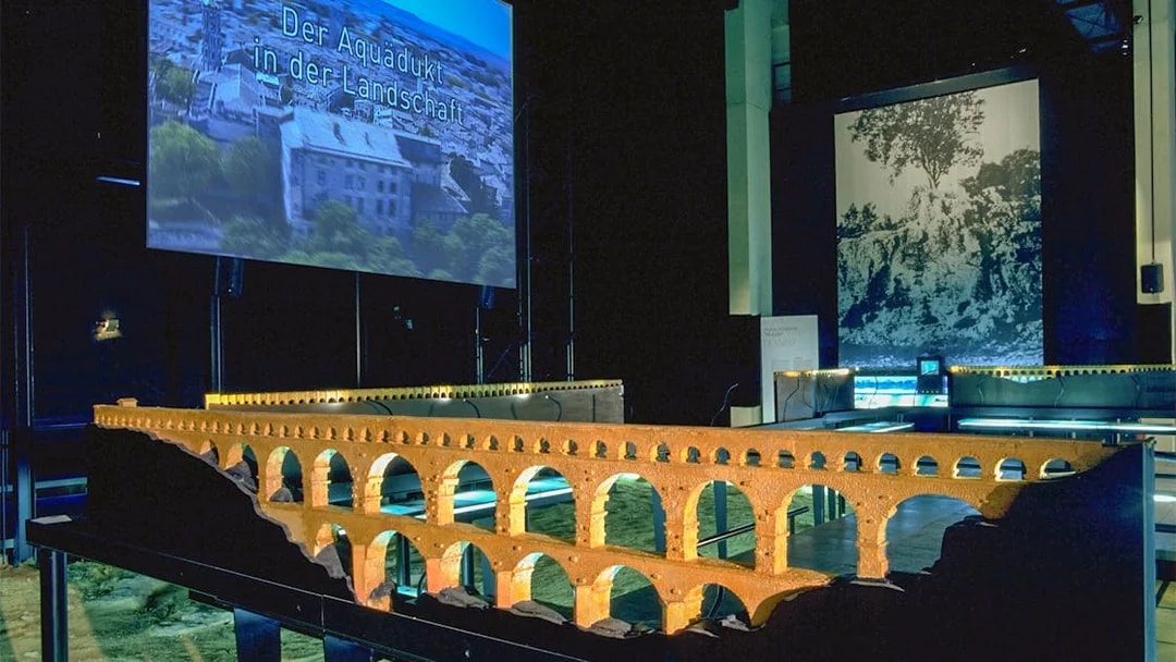 Model of Pont du Gard aqueduct in museum exhibit with cinema screen in background.