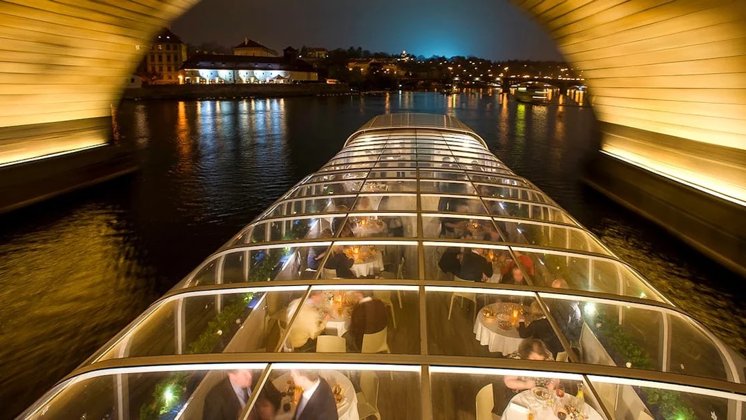Open-top glass boat on Prague dinner cruise with live music, passing under a lit bridge at night.