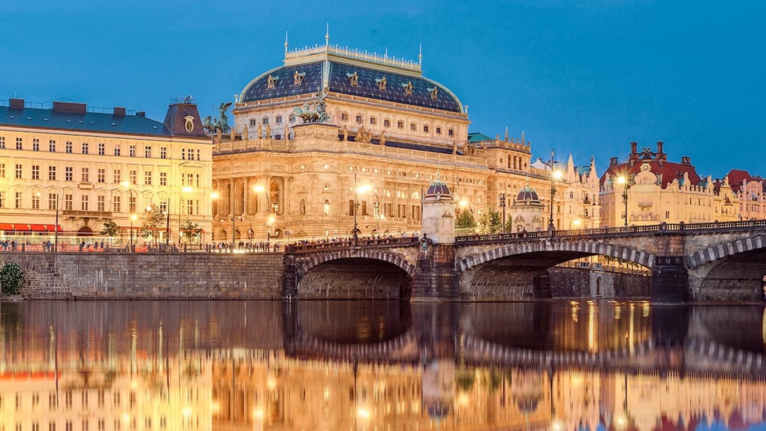Prague's National Theatre and bridge illuminated at night, viewed from a river.