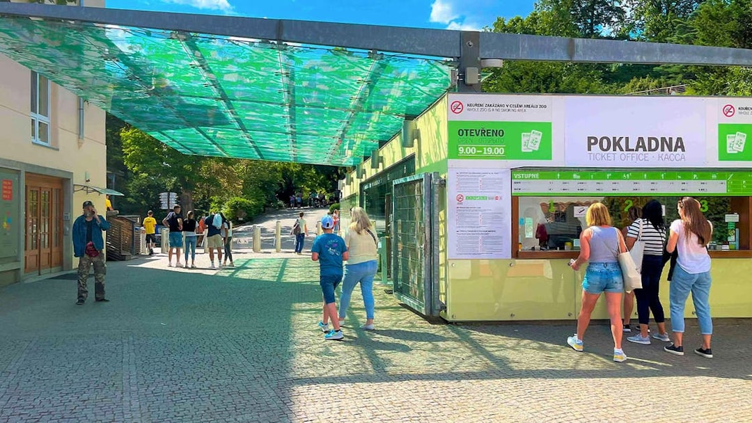 Visitors at Prague Zoo ticket office under a green canopy.