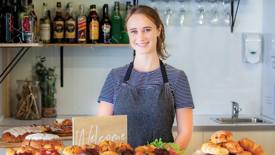 Bakery display with pastries and muffins on a counter, staff member smiling behind.
