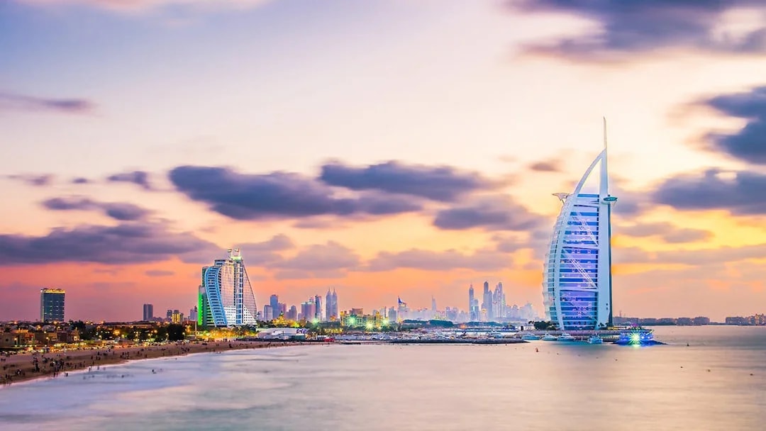 Burj Al Arab illuminated at evening with Dubai skyline in the background.