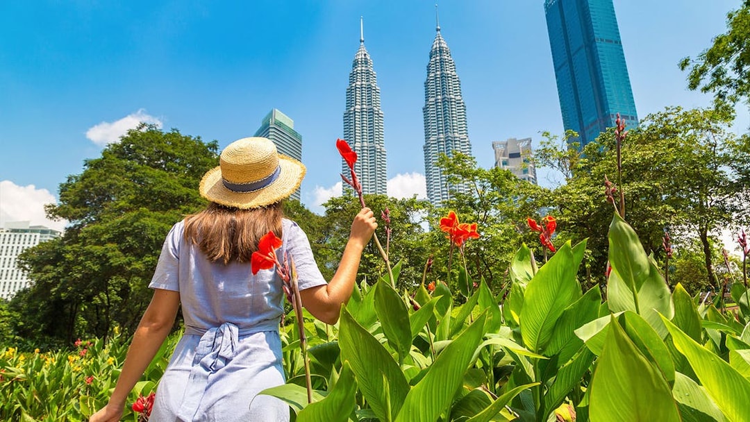 Person in a hat holding flowers near Petronas Towers, Kuala Lumpur.