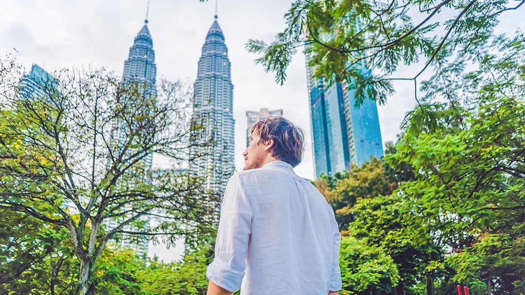 Person standing near Petronas Towers in Kuala Lumpur, surrounded by trees.