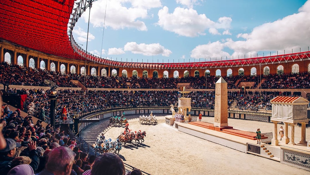 Chariot race in Puy du Fou arena with large audience and ancient Roman architecture.