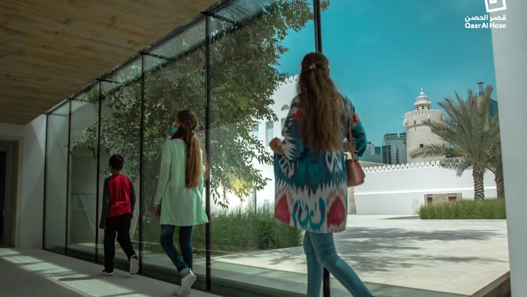 Visitors walking through glass corridor with view of Qasr Al Hosn tower in Abu Dhabi.