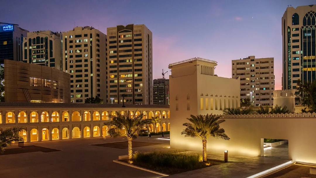 Qasr Al Hosn illuminated at dusk with modern buildings in Abu Dhabi background.