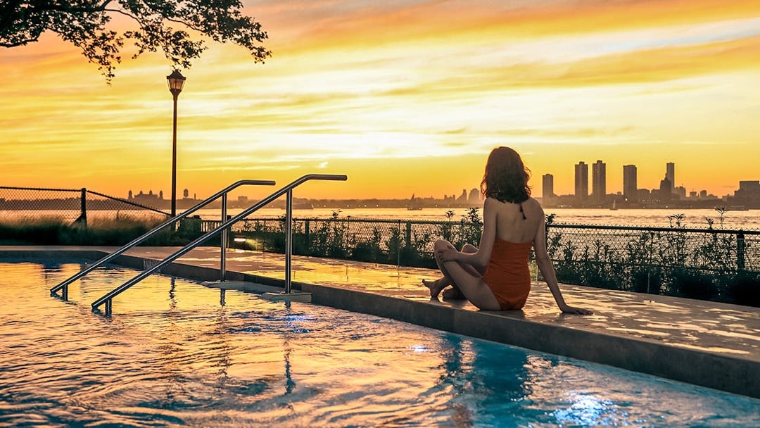 Person sitting by a pool at sunset with New York City skyline in the background.