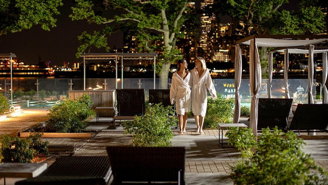 Couple in robes walking at night in QC New York spa garden with city skyline.