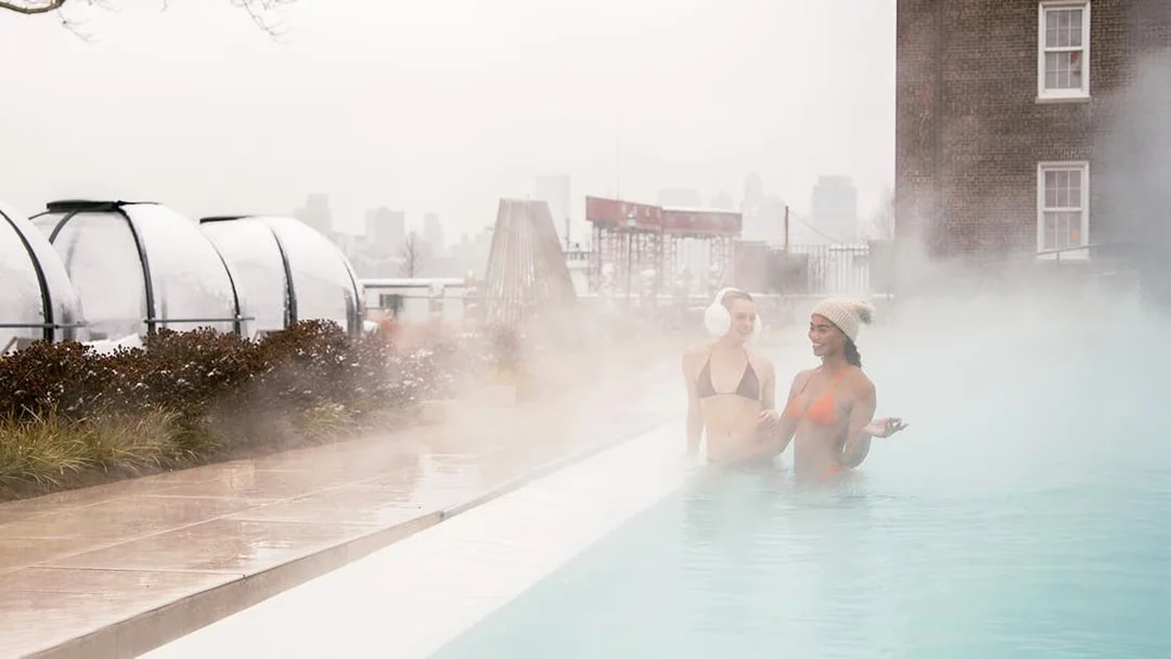 People enjoying a heated outdoor pool at QC New York with city skyline in the background.