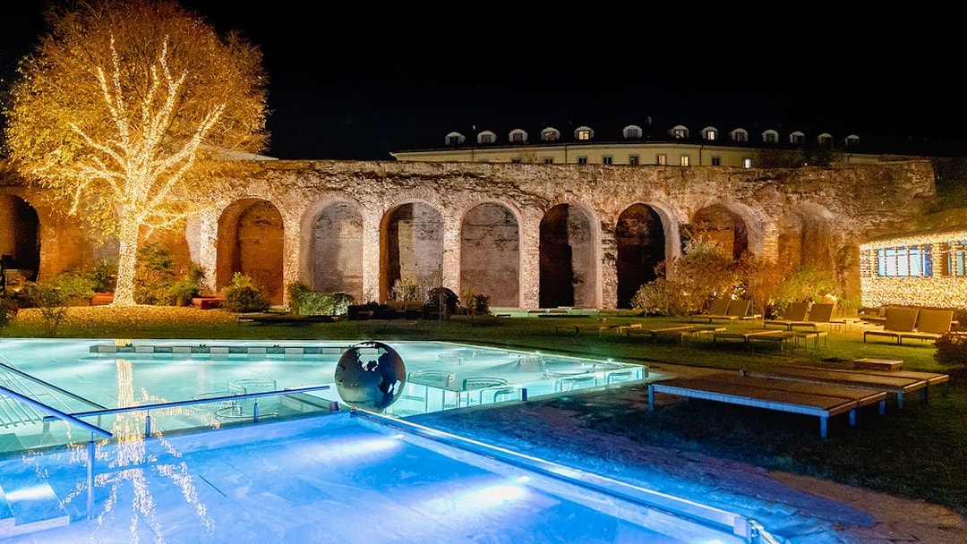 QC Terme Milano Resort & Spa outdoor pool with illuminated tree and ancient arches at night.