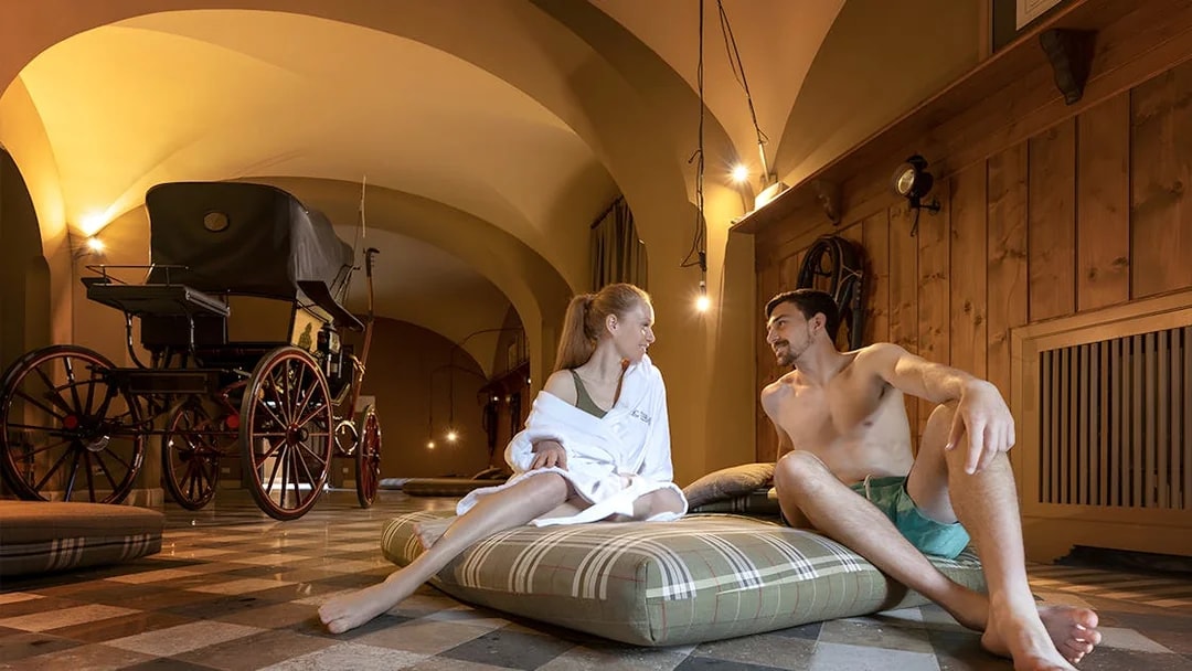 Couple relaxing on cushions in QC Terme Roma spa with vintage carriage in background.