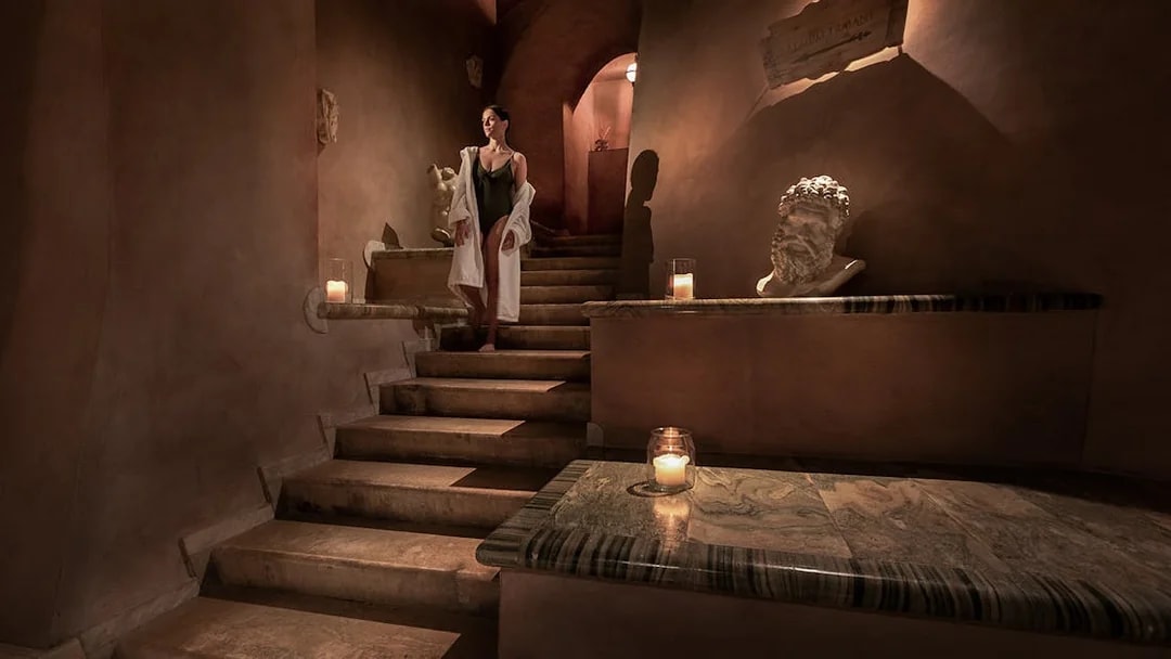 Woman in robe descending candlelit stone stairs at QC Terme Roma spa.