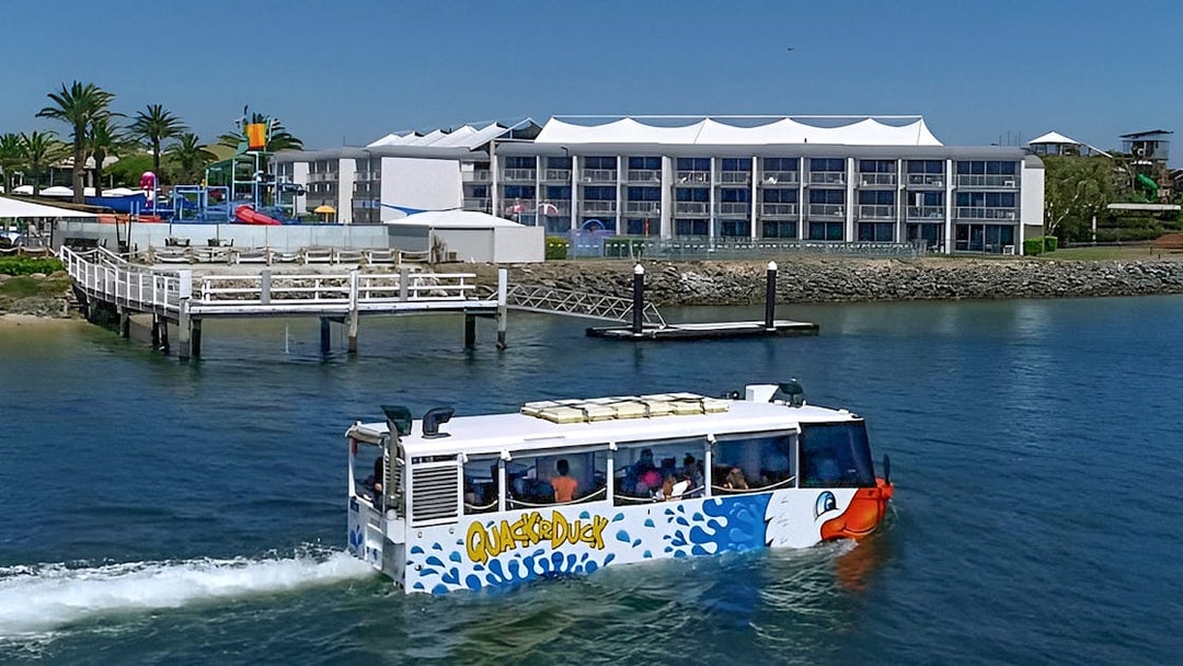 Quack'R Duck amphibious vehicle cruising on Gold Coast waterway near a waterfront hotel.