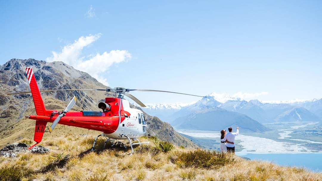 Couple by helicopter at Queenstown viewpoint overlooking mountains and river valley.