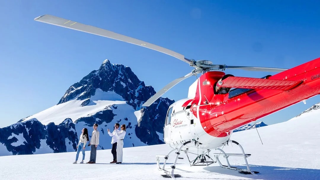 Three people beside a red and white helicopter on a snowy peak in Milford Sound.