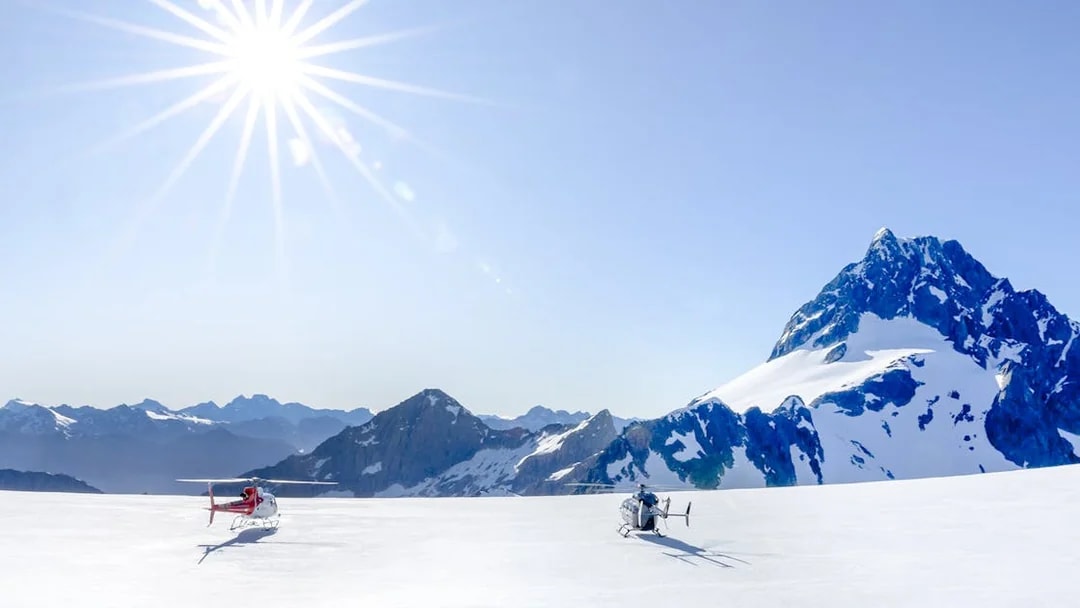 Two helicopters on a snow-covered glacier in the Southern Alps, Queenstown, under a bright sun.