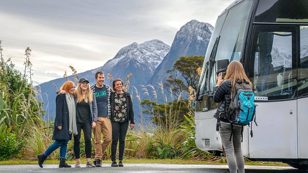 Group posing for a photo with a tour bus and snow-capped mountains in Milford Sound, New Zealand.