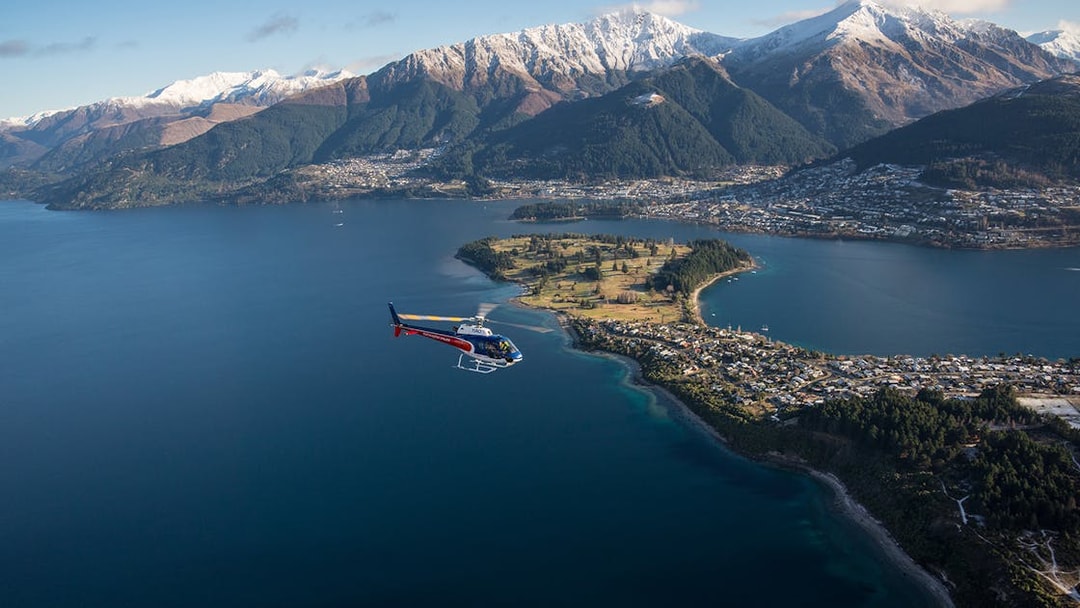 Helicopter flying over Lake Wakatipu with Queenstown and snow-capped mountains in view.
