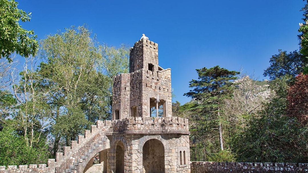 Stone tower with spiral staircase at Quinta da Regaleira Palace, Sintra, surrounded by trees.