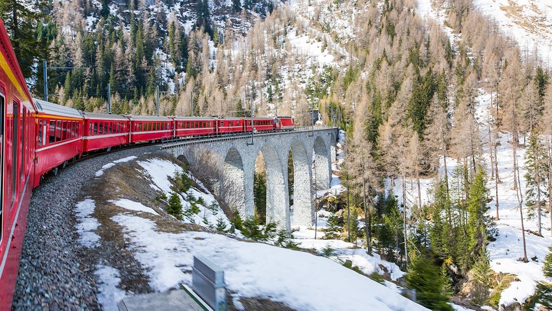 Bernina train crossing a stone viaduct in a snowy alpine landscape.