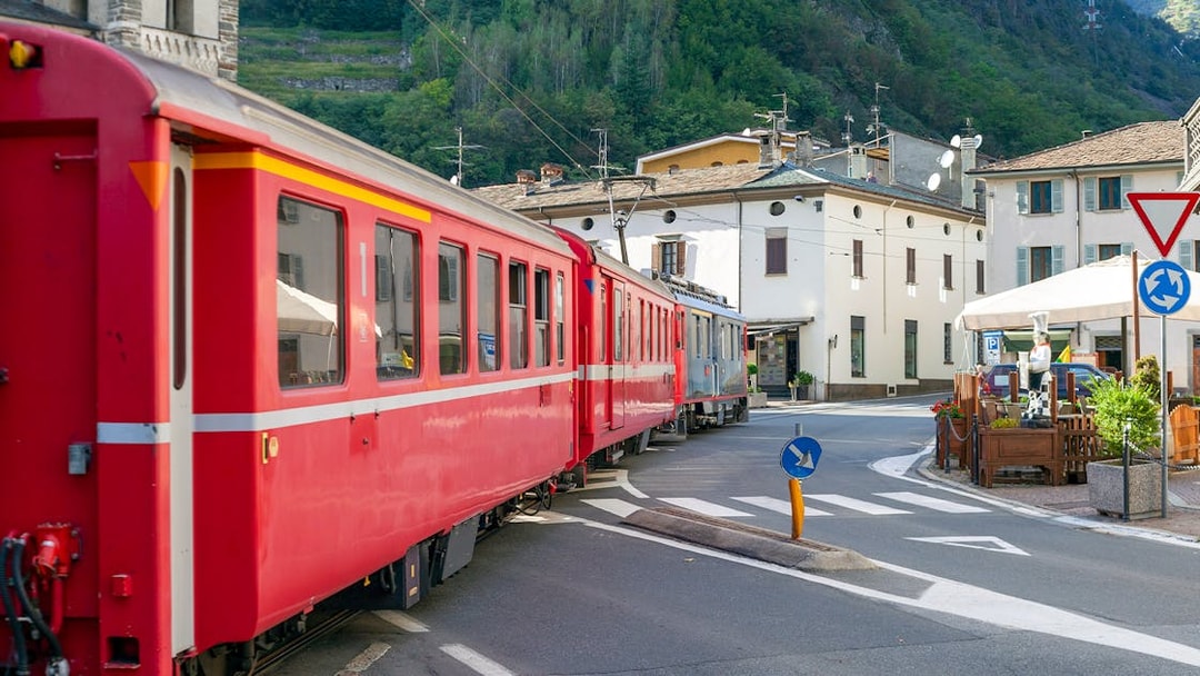 Bernina train passing through Tirano city street, Italy, with buildings and mountains in the background.