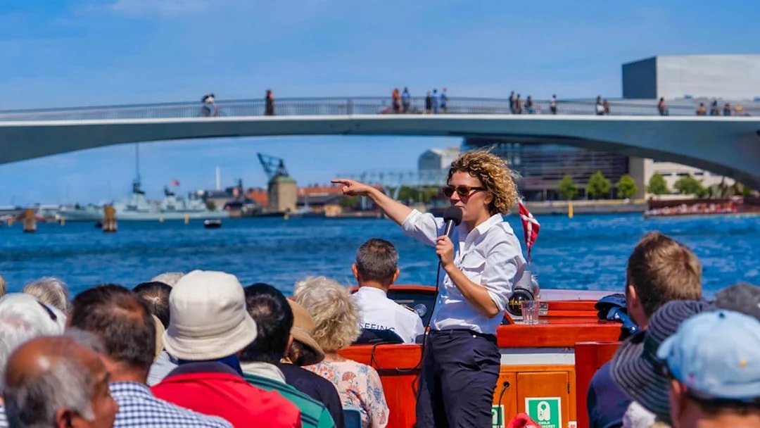 Tour guide speaking to passengers on a boat during a Copenhagen canal tour with a bridge in the background.