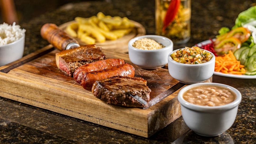 Brazilian barbecue with grilled meats, rice, beans, and sides on a wooden board.